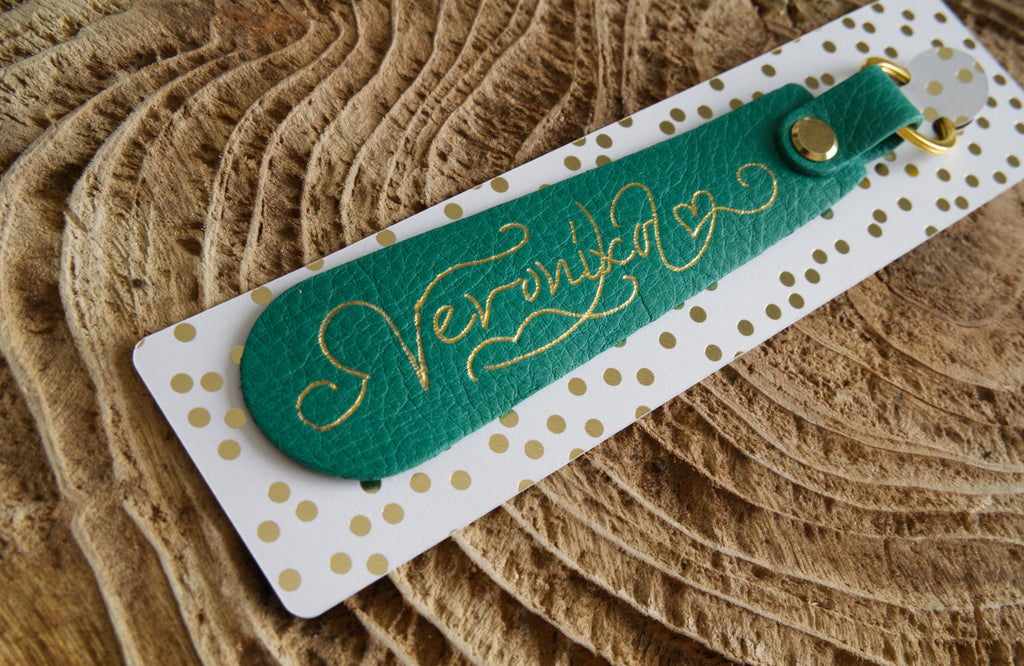 Green leather bookmark with gold text on a white card with gold dots, placed on a textured brown surface.