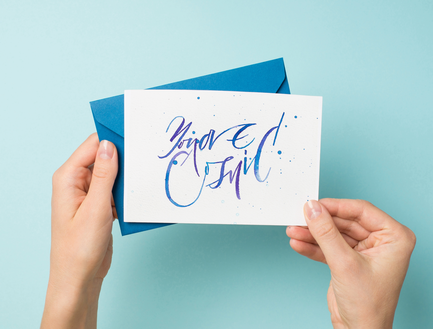 A person holding a watercolor greeting card with the text 'You are Cosmic' written in calligraphy, featuring blue and purple colors on a white card with a blue envelope.