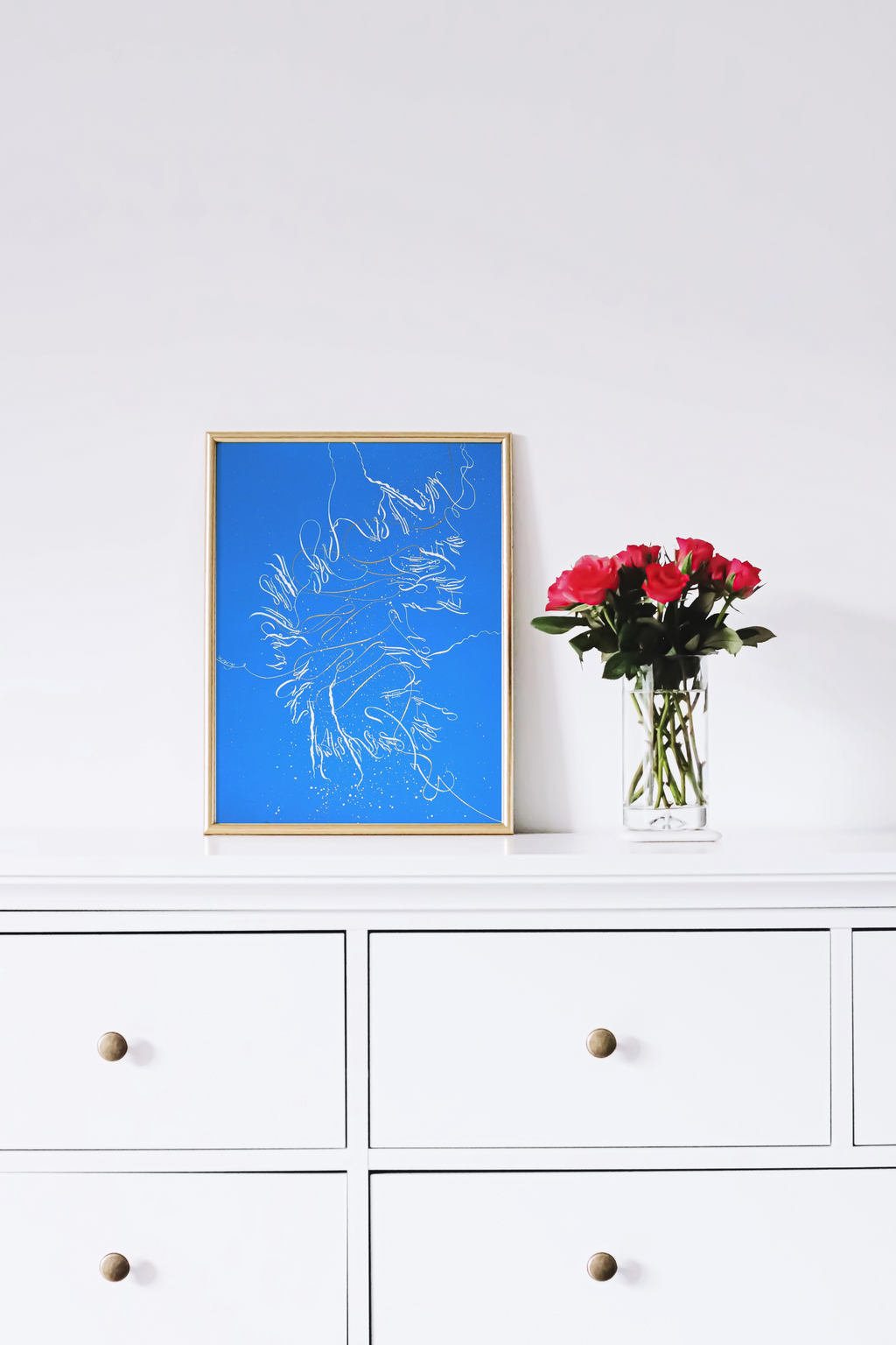 Framed blue artwork on a white wall above a white cabinet with a vase of red flowers.