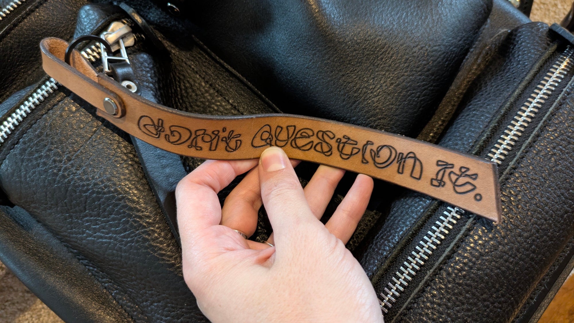 Hand holding a leather strap with 'Don't Question It' text on a black leather bag.